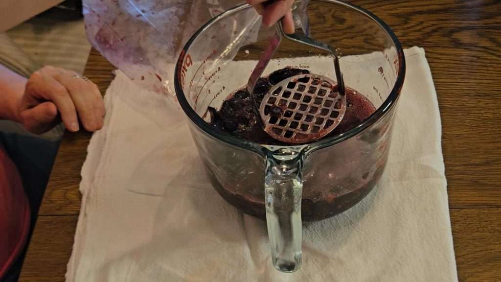 Blueberries in a large, Pyrex measuring cup, being mashed with a potato masher. Mashing the berries is an important step when canning blueberry jam.