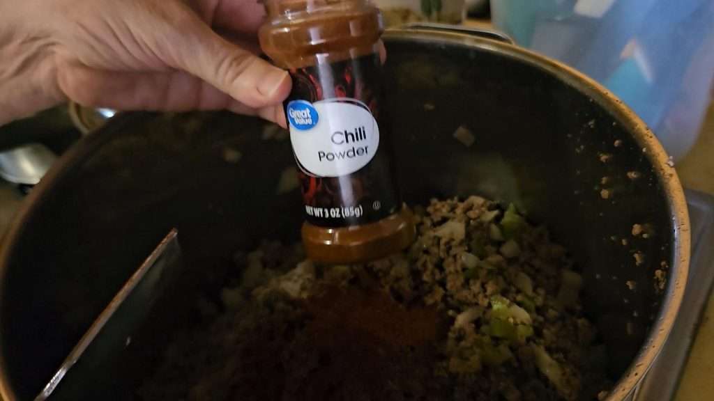 Holding up a spice jar of chili powder over the pot filled with cooked ground beef, bell peppers, and onions.