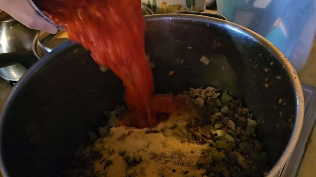 pouring canned crushed tomatoes into a pot with the ground beef, onions, peppers, and spices. 