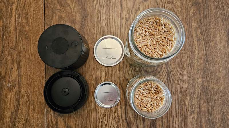 The LOVE Moment Electric Mason Jar Vacuum Sealer next to two lids and two mason jars filled with wheat berries. 
