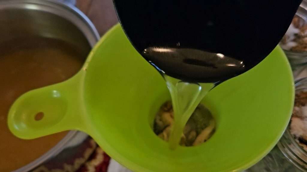 A ladle pouring broth through a canning funnel into a canning jar