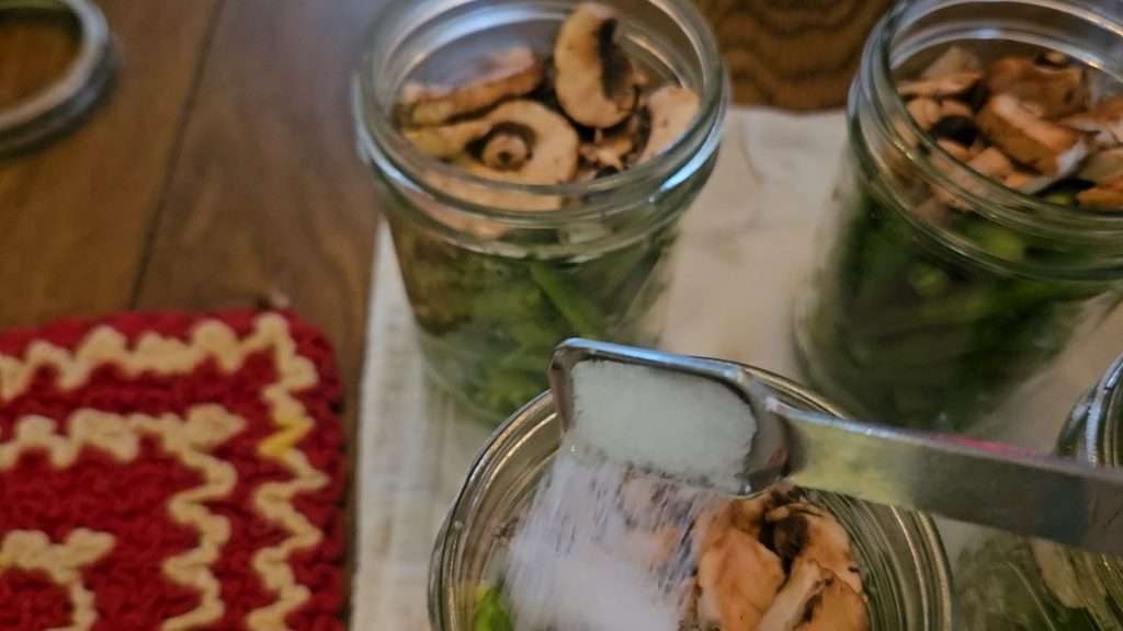 Pouring salt into a canning jar filled with green beans and mushrooms.