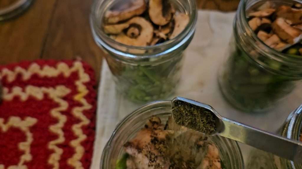 Pouring thyme leaves into a canning jar filled with green beans, mushrooms, salt, and black pepper.