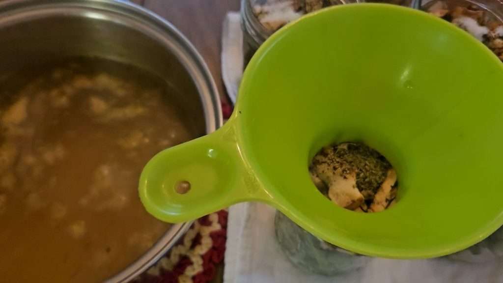 A pot with hot chicken broth sitting next to a canning jar with a canning funnel on top.