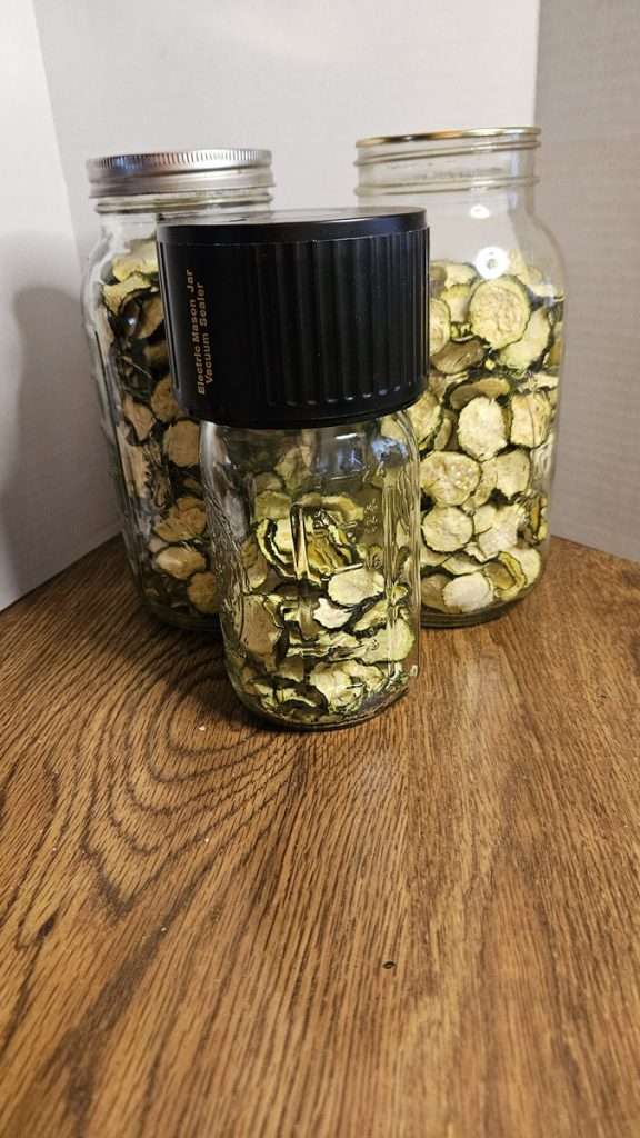Two large jars and one smaller jar filled with dehydrated zucchini, with a vacuum sealer on the small jar. 