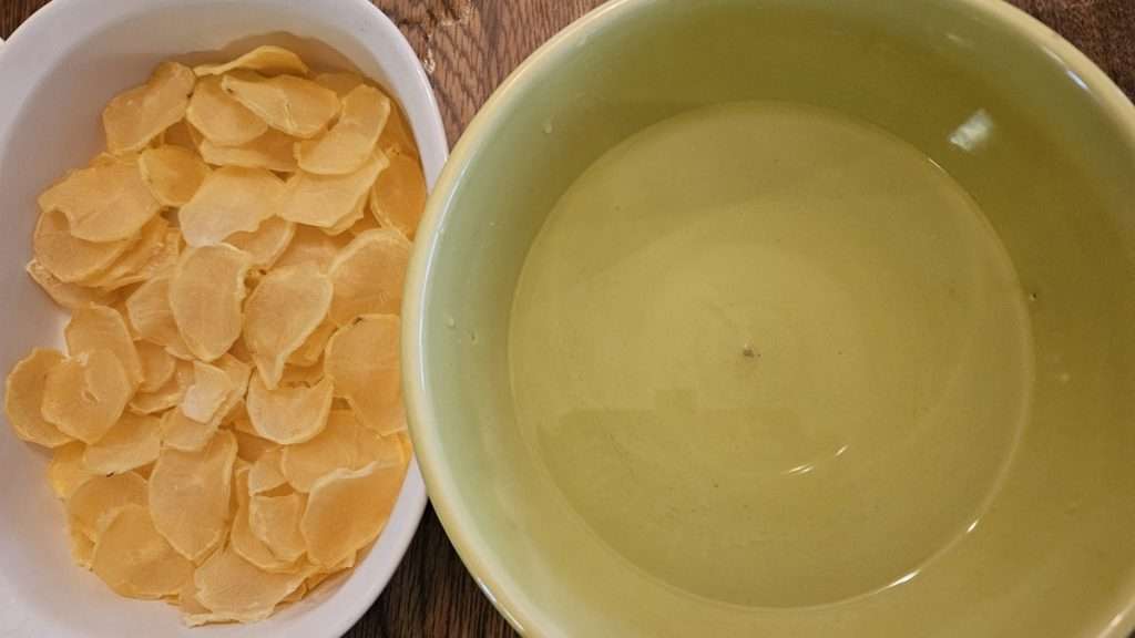 On the left dehydrated sliced potatoes in a casserole dish, and on the right is a bowl with water.