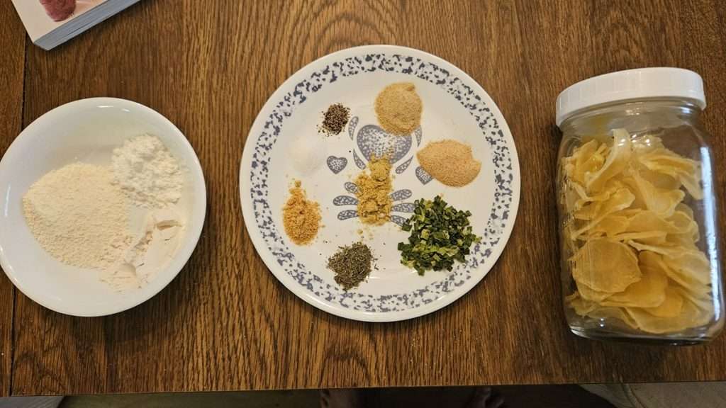 This picture has a close up of the ingredients used in dehydrated scalloped potatoes, including a bowl with powdered milk, flour and cornstarch, a plate with seasonings, and a jar filled with dehydrated sliced potatoes.