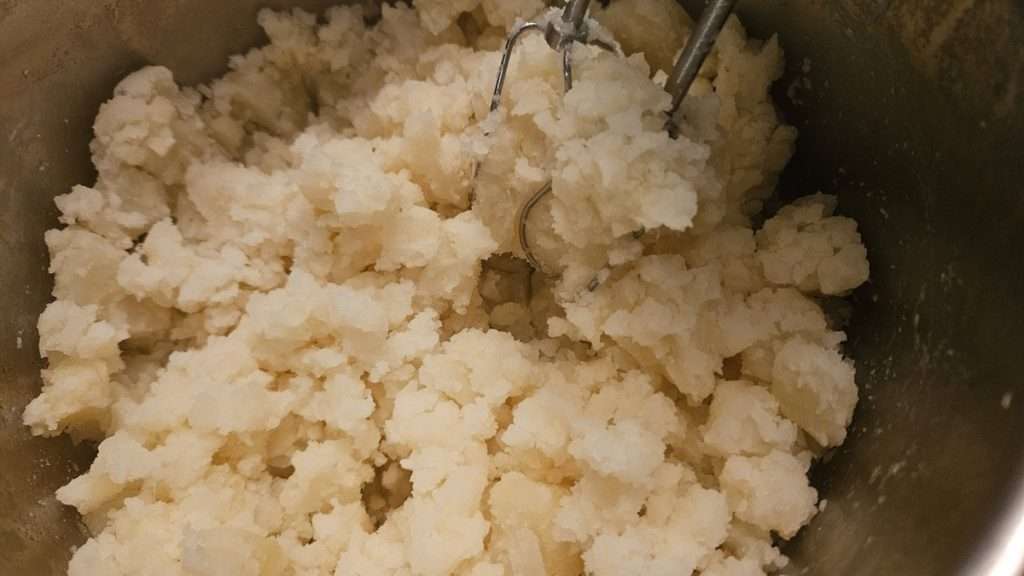 Using a hand mixer to mash boiled potatoes in a large stainless steel pot.