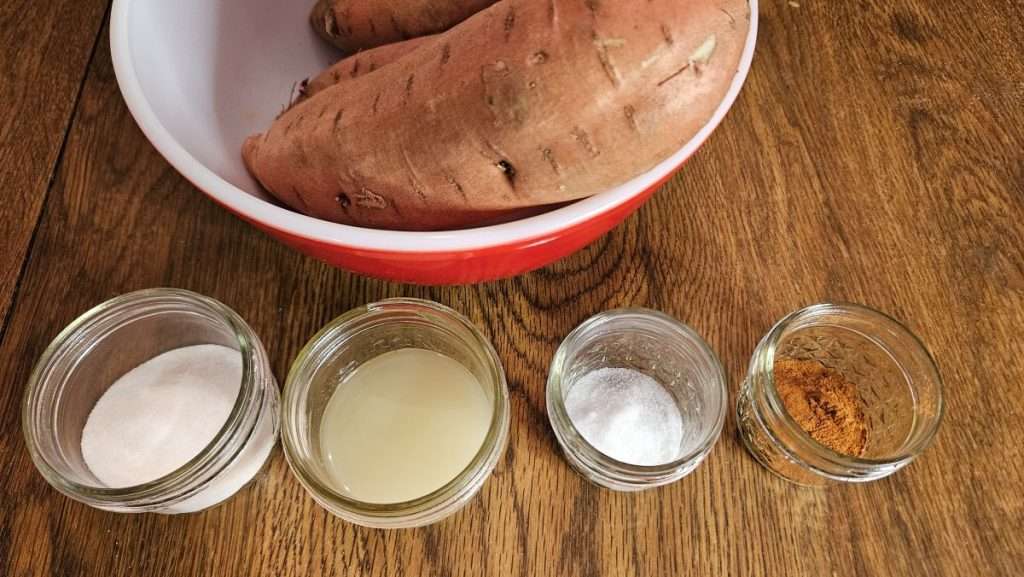 Mason jars filled with sugar, lemon juice, salt, and cinnamon in front of a bowl of raw sweet potatoes